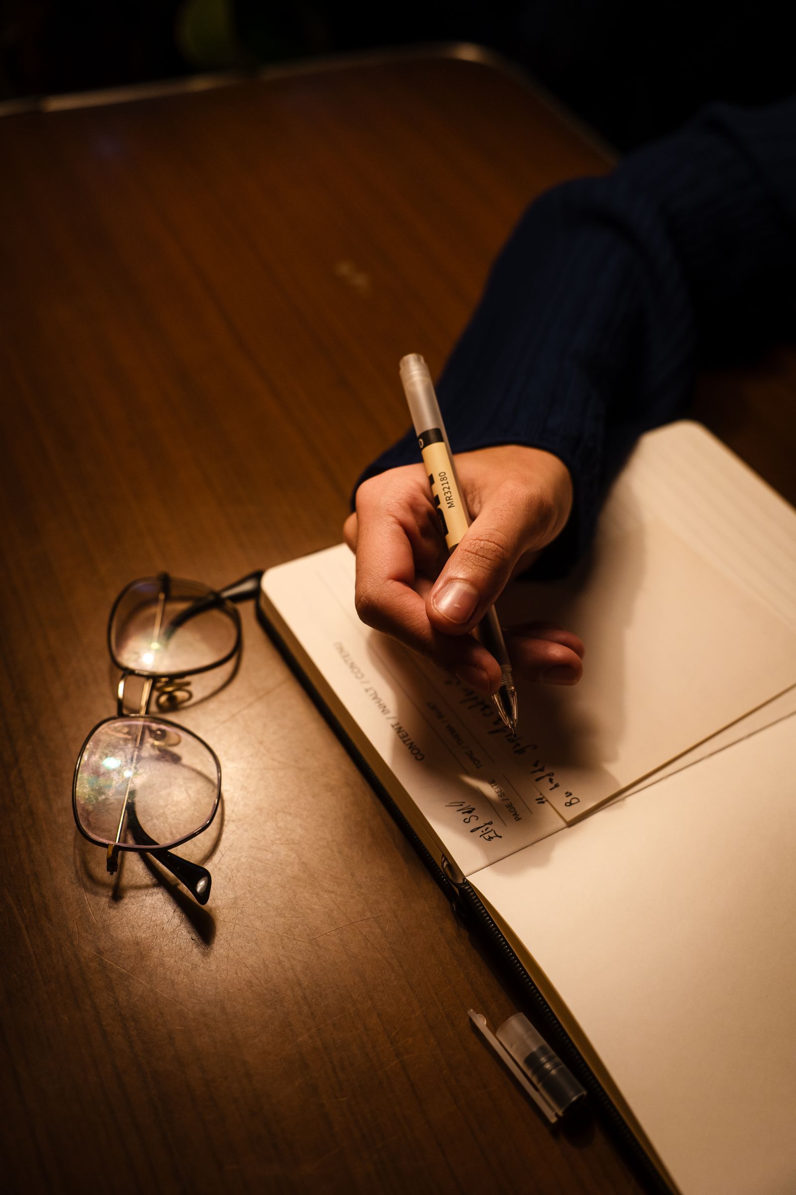 A hand writing in a notebook under a warm lamp